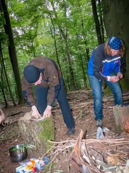 Zwei Personen bearbeiten Holzstücke im Wald, während ein Kochtopf und Zunder sichtbar sind