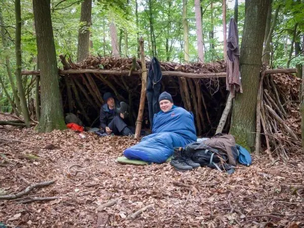 Zwei Personen sitzen vor einer selbstgebauten Laubhütte im Wald