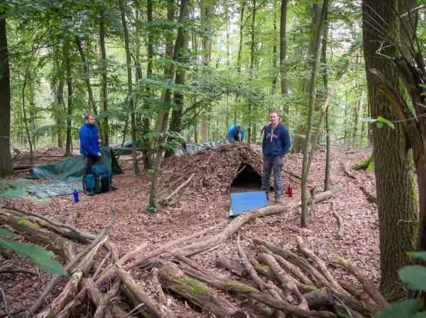 Zwei Personen stehen neben einer selbstgebauten Laubhütte im Wald