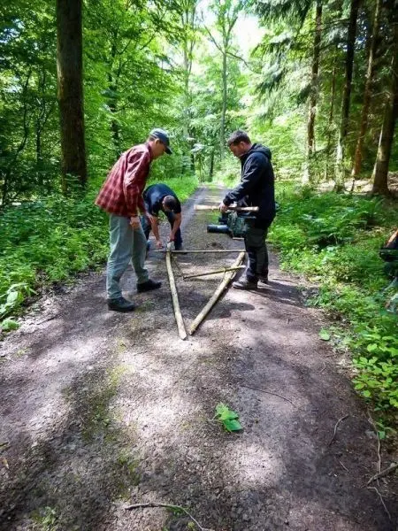 Drei Personen bauen eine selbstgebaute Konstruktion aus Ästen auf einem Waldweg