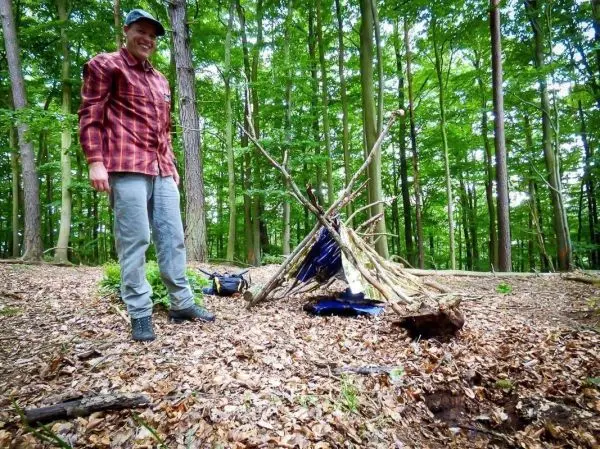Person steht neben einer selbstgebauten Unterkunft im Wald