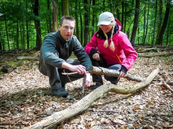 Zwei Personen arbeiten an einer Holzstruktur im Wald, eine Person hält einen Stock