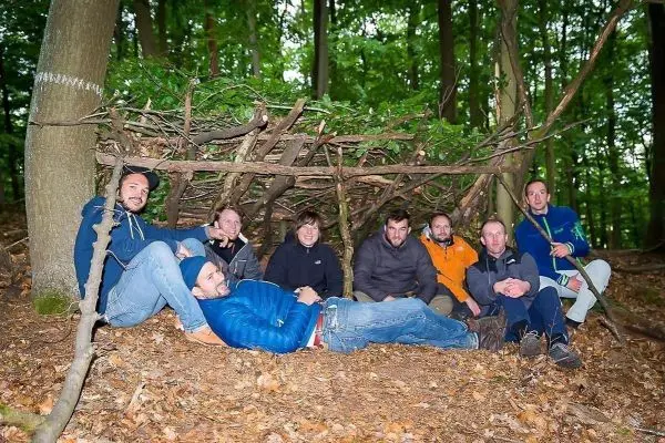 Gruppe sitzt vor einer selbstgebauten Tarp-Notunterkunft im Wald