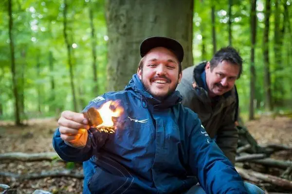 Mann hält brennendes Zundernest in der Hand, im Hintergrund ein weiterer Mann