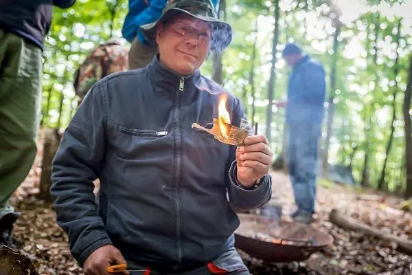 Mann hält brennendes Zundernest in der Hand, im Hintergrund rauchendes Feuer
