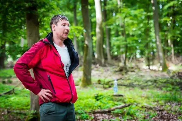 Mann in roter Jacke steht im Wald mit Händen an der Hüfte