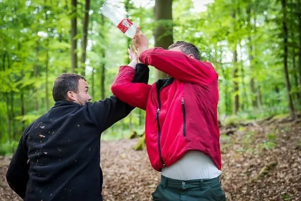 Zwei Personen in einem Wald, eine Person hebt eine Flasche über den Kopf