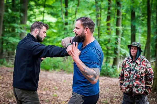 Zwei Personen üben eine Selbstverteidigungstechnik im Wald, während eine dritte Person zuschaut