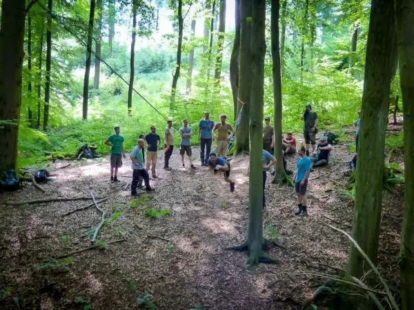 Gruppe steht im Wald, einige Personen überqueren eine selbstgebaute Seilbrücke