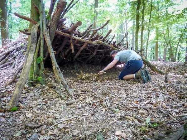 Mann kniet vor einer selbstgebauten Laubhütte und bereitet Zunder vor