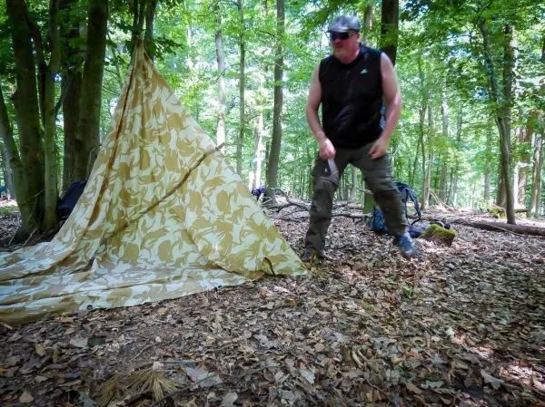 Mann richtet Tarp-Notunterkunft im Wald auf, umgeben von Laub und Bäumen