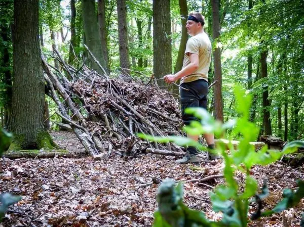 Mann steht neben einer selbstgebauten Laubhütte aus Ästen und Blättern