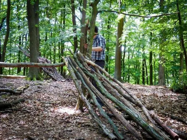 Mann stützt sich an einen Baum neben einer selbstgebauten Holzunterkunft