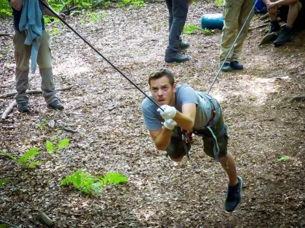 Mann überquert selbstgebaute Seilbrücke im Wald, gesichert mit Seil und Karabiner