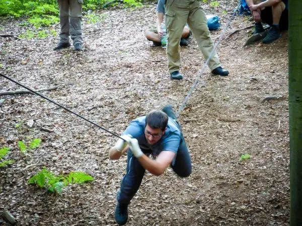 Mann überquert selbstgebaute Seilbrücke im Wald, zieht sich mit beiden Händen hoch