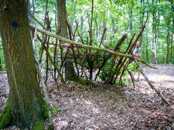 Person steht neben einer selbstgebauten Struktur aus Ästen im Wald