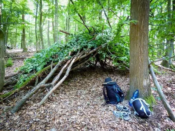 Selbstgebaute Tarp-Notunterkunft aus Ästen und Blättern neben Rucksack und Wasserflasche