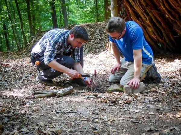 Zwei Personen bereiten Zunder auf dem Boden vor und nutzen Holzstücke