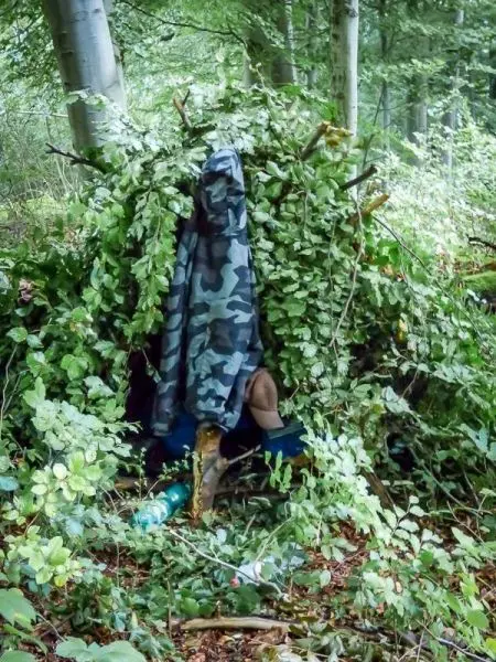 Person sitzt in einer selbstgebauten Unterkunft aus Ästen und Blättern