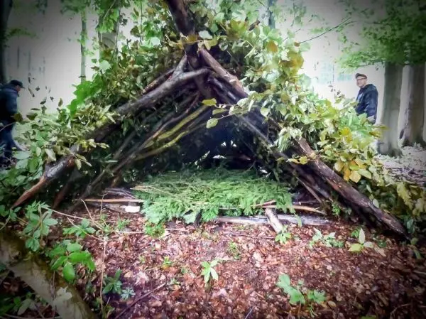 Person steht neben einer selbstgebauten Unterkunft aus Ästen und Blättern