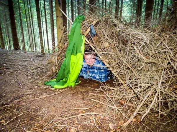 Tarp-Notunterkunft aus Ästen und Zweigen im Wald, Person schaut heraus