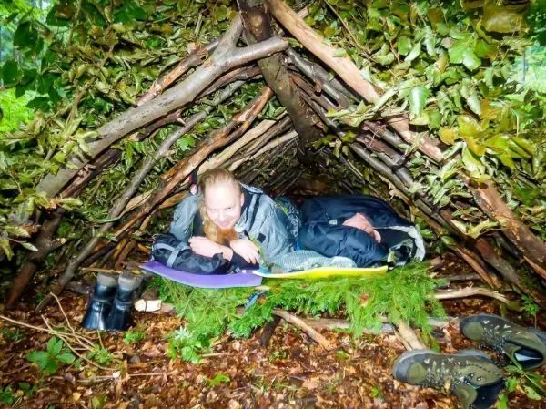 Zwei Personen liegen in einer selbstgebauten Laubhütte auf einer Unterlage aus Tannenzweigen