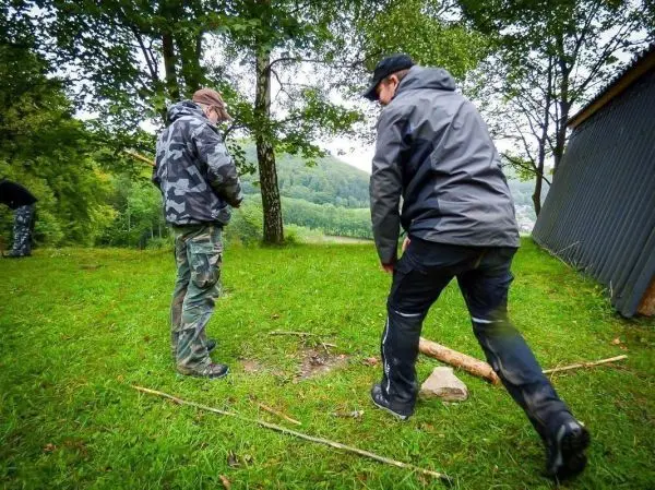 Zwei Personen stehen auf einer Wiese und bearbeiten einen Holzstock beim Überlebenstraining
