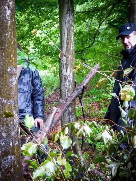 Zwei Personen stehen zwischen Bäumen, eine selbstgebaute Konstruktion aus Ästen ist sichtbar