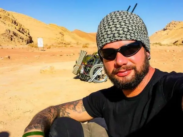 Ein Mann mit Sonnenbrille und Kopfbedeckung sitzt im Wüstensand neben einem Fahrrad