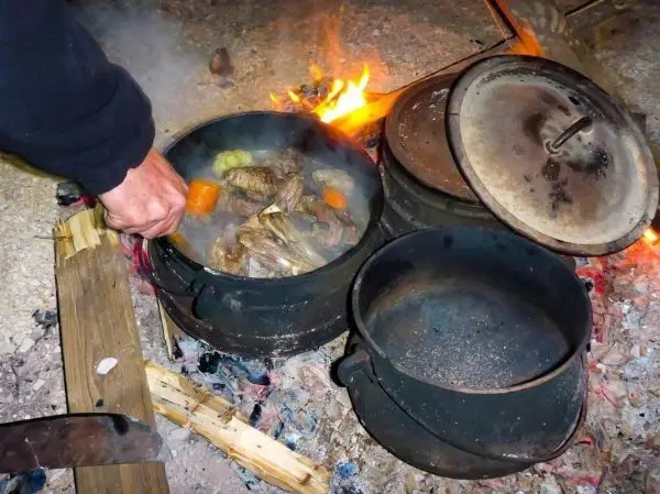 Hand greift in einen Kessel über offenem Feuer mit Gemüse und Fleisch