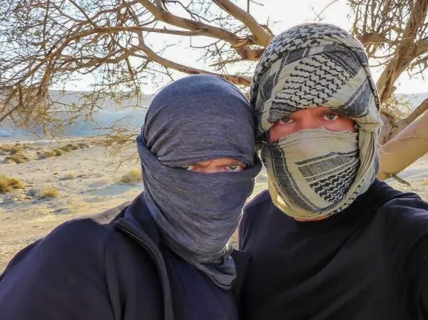 Zwei Personen mit Tüchern um den Kopf stehen vor einem Baum in einer Wüstenlandschaft