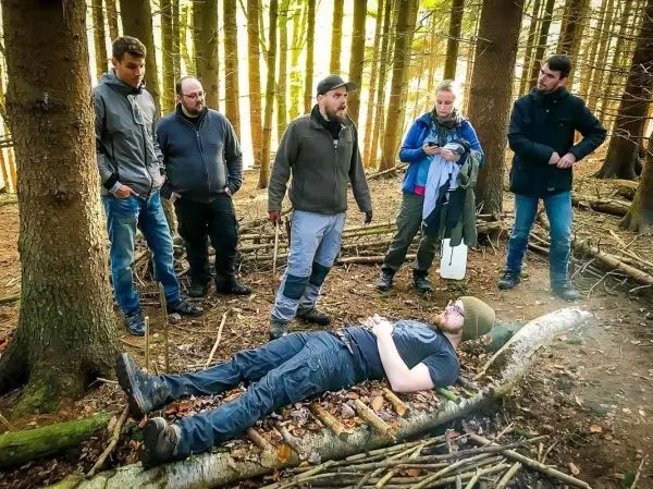 Gruppe steht um liegende Person auf improvisierter Trage aus Ästen im Wald