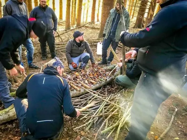 Gruppierung von Personen um eine Holzstruktur, die mit Ästen und Laub belegt ist