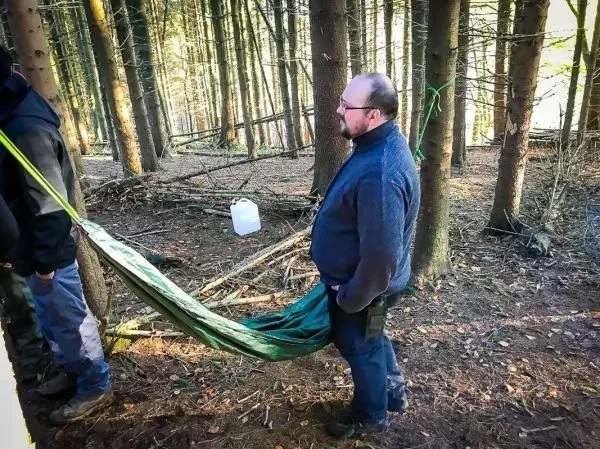Mann steht neben einer hängenden Tarp-Notunterkunft zwischen Bäumen