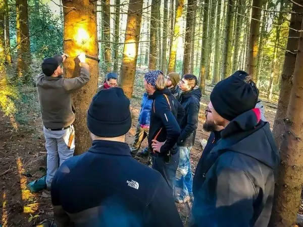 Person erklärt Technik an Baum, Gruppe hört aufmerksam zu beim Überlebenstraining