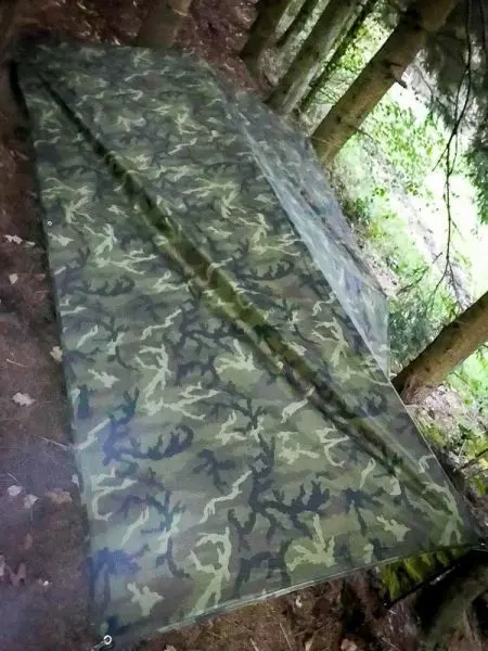 Tarp-Notunterkunft aus Tarnmaterial zwischen Bäumen im Wald auf dem Boden