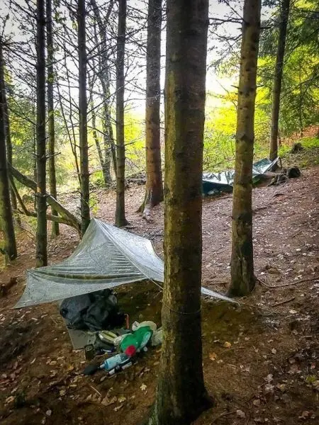 Tarp-Notunterkunft zwischen Bäumen mit Ausrüstung und Rucksack im Vordergrund