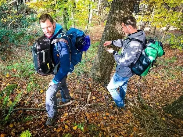 Zwei Personen mit Rucksäcken bewegen sich durch einen Wald mit herbstlichen Blättern