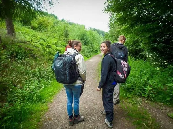 Drei Wandernde mit Rucksäcken auf einem schmalen Weg zwischen Bäumen und Sträuchern