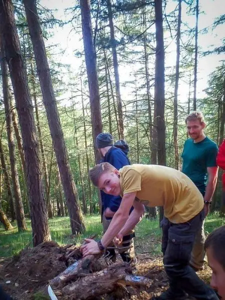Gruppe arbeitet an einer Holzstruktur im Wald, einige Personen stehen im Hintergrund