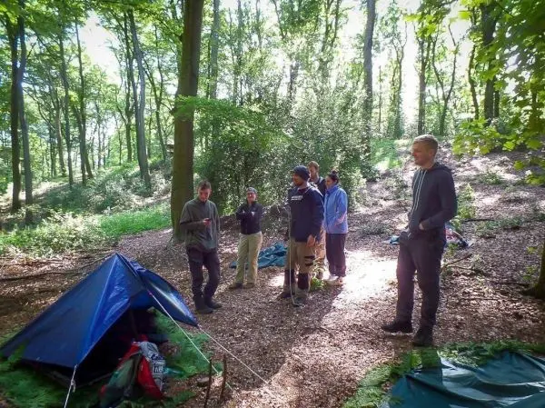 Gruppe steht um ein Zelt im Wald, einige halten Ausrüstung in der Hand