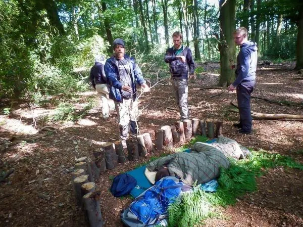 Gruppe steht um eine improvisierte Notunterkunft aus Holzstämmen und Decken