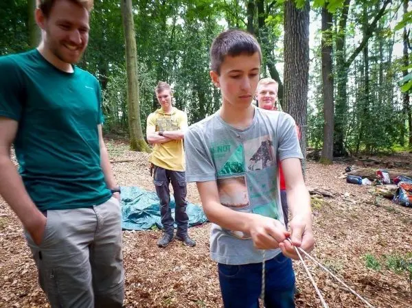 Junge knotet Seil, während andere zuschauen und im Wald stehen