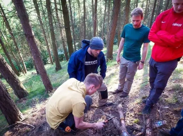 Junger Mann bearbeitet Holzstück mit einem Messer, während andere zuschauen