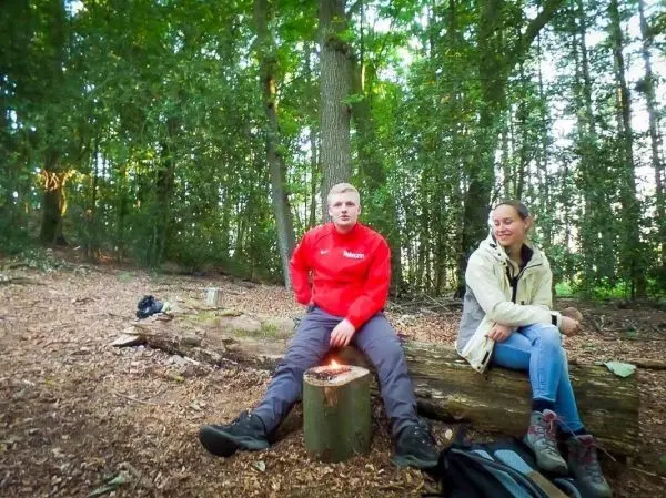 Kleines Bodenfeuer auf einem Baumstumpf im Wald, zwei Personen sitzen auf einem Holzstamm