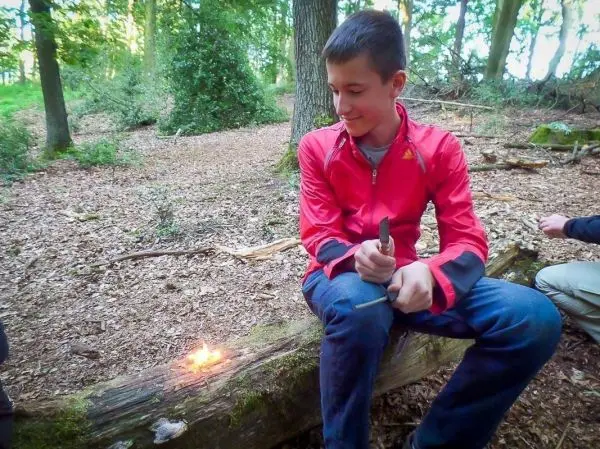 Kleines Bodenfeuer brennt auf einem Baumstamm, während jemand mit einem Messer sitzt