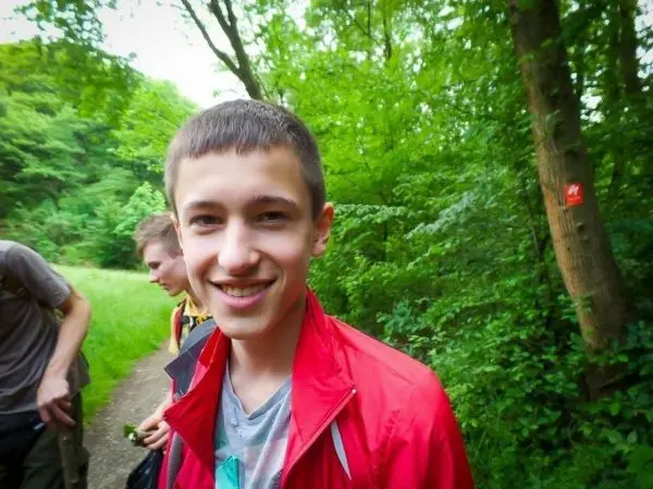 Lächelnder Junge in roter Jacke steht auf einem Waldweg zwischen Bäumen