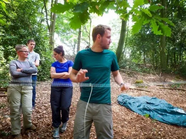 Mann zeigt mit Seil auf eine Gruppe im Wald, im Hintergrund liegt eine Tarp-Notunterkunft
