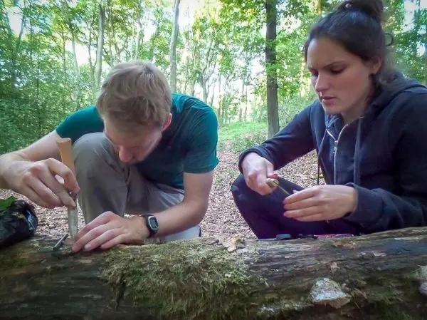 Messer bearbeitet Holzstück im Wald, während eine Person Zunder vorbereitet