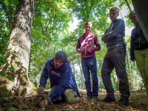Person bereitet Feuerstelle im Wald vor, Gruppe schaut zu beim Überlebenstraining